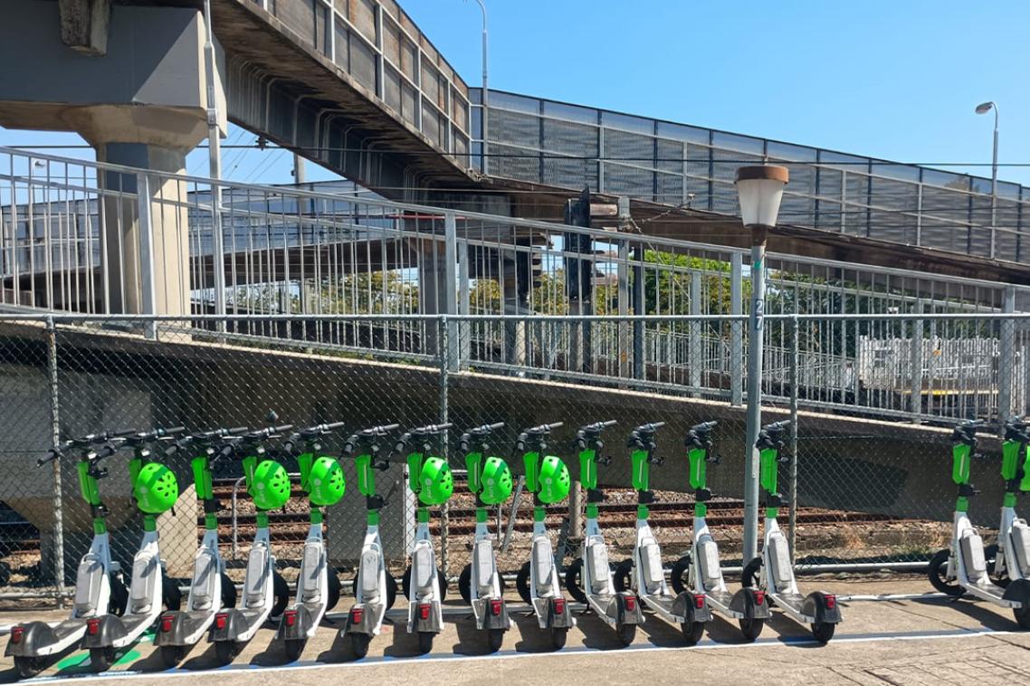A row of white and green rideshare e-scooters lined up at an urban train station, with concrete ramps and walkways in the background.